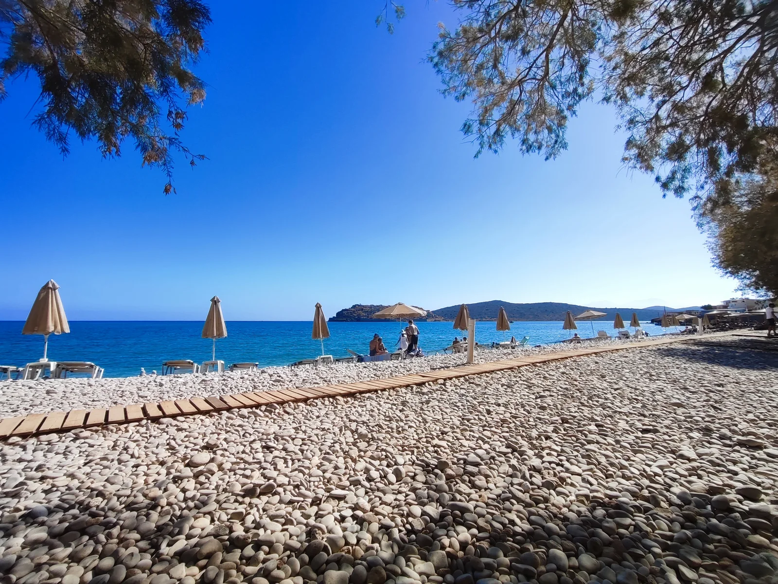 Pebble shore at the public organised section of Plaka beach, with umbrellas and Spinalonga beyond.