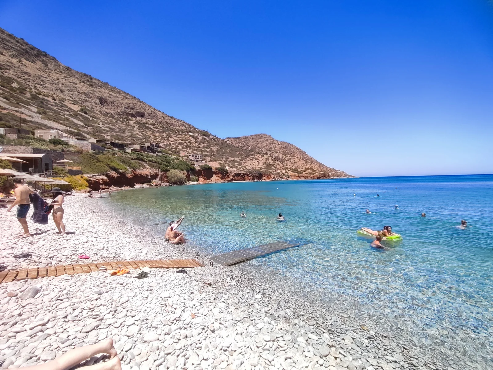 Pebble shore and swimmers at Plaka, with the coast beyond.