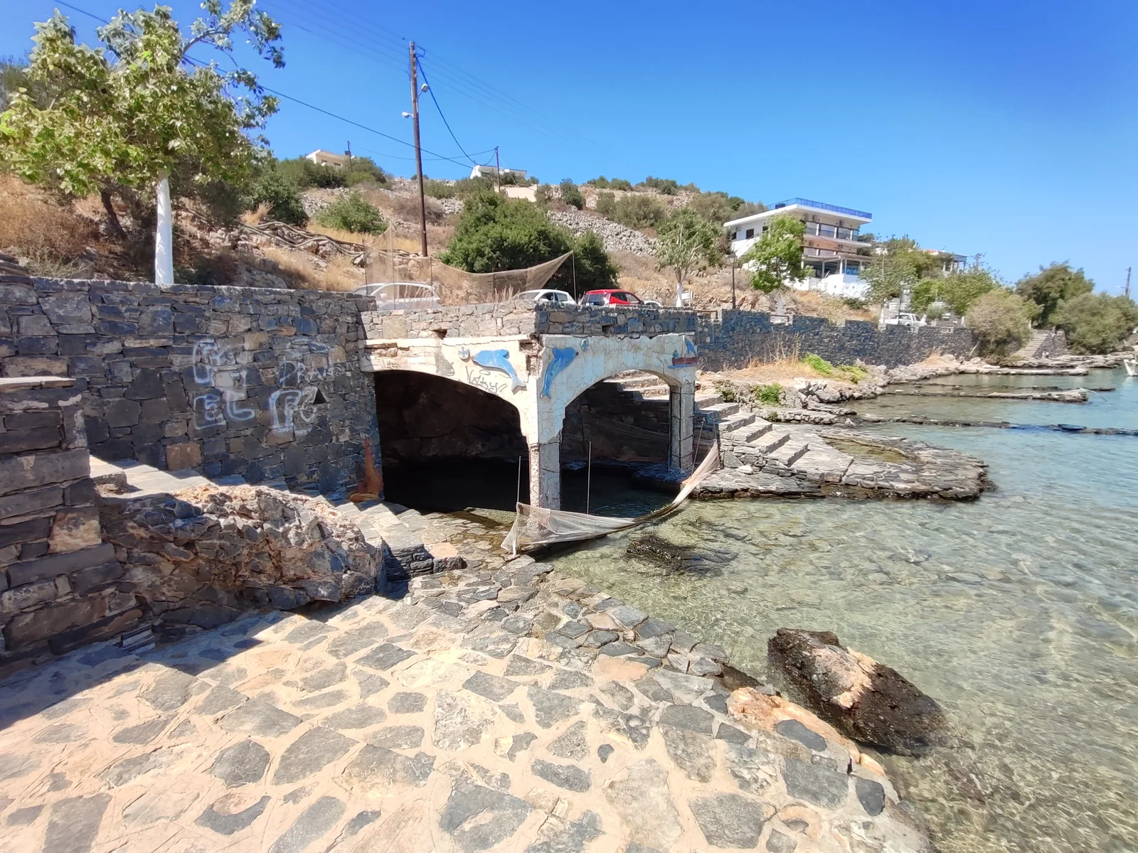Stone-and-concrete enclosure beside broad stone steps and shallow seawater, with hillside buildings behind.