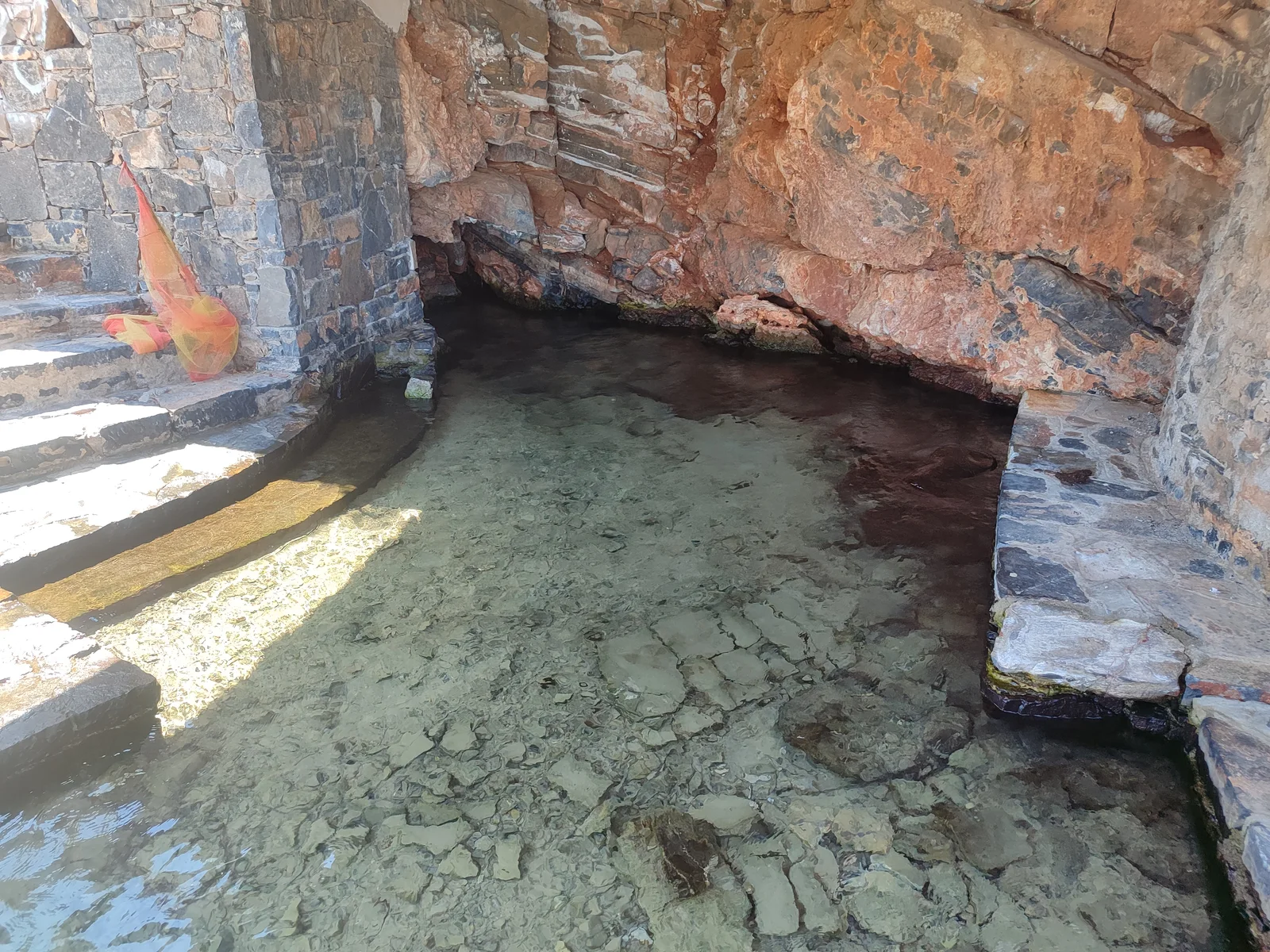 Clear shallow water against layered red and grey rock inside the covered Mavrikiano outlet area.