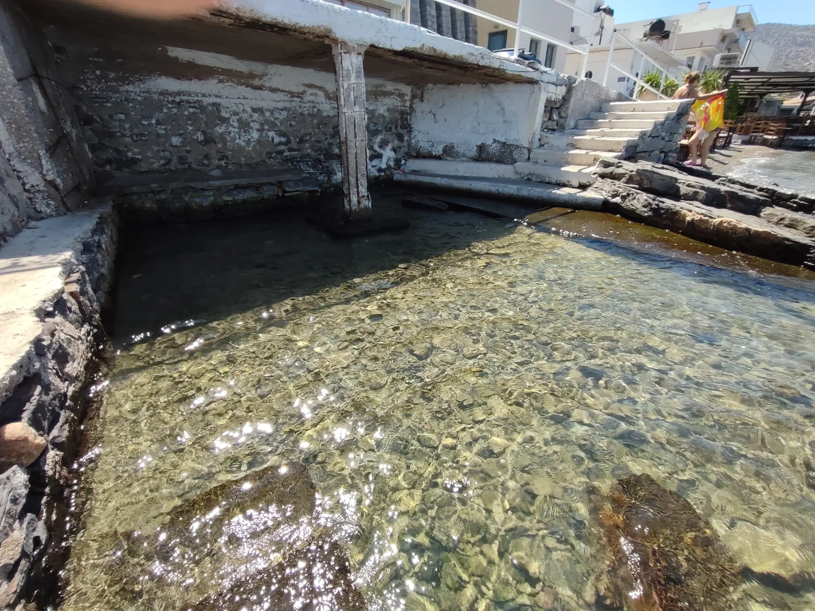 Shallow clear water beside the shaded Schisma waterfront enclosure and stone steps.