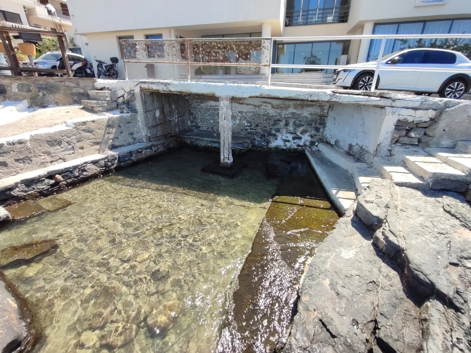 Clear shallow water and a stone-edged channel beneath a low concrete roadside structure, with steps at the right.