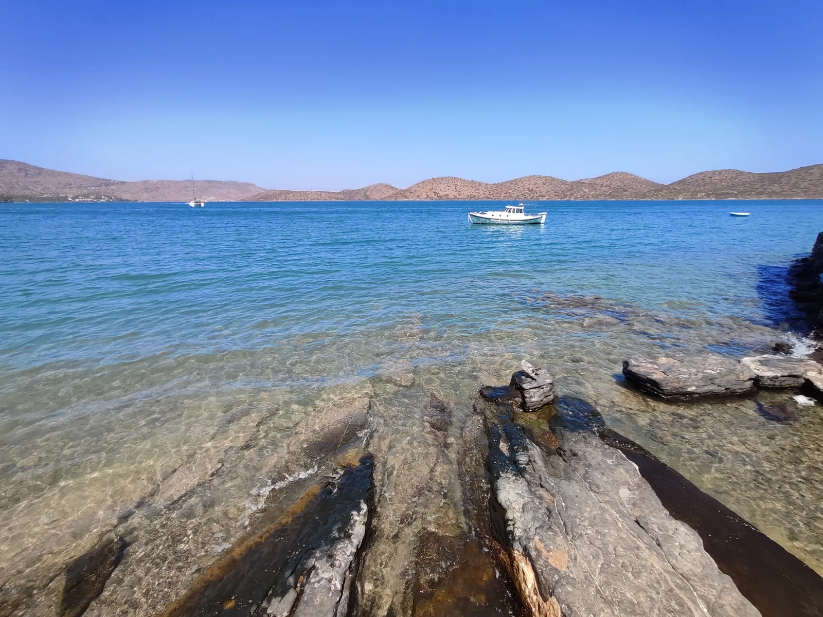 Rocky shallows opening into Elounda Bay, with a small boat and low hills across the water.
